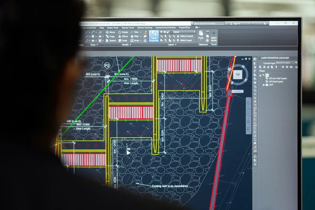 Over-the-shoulder view of a monitor showing a dimensioned 2D CAD plan with coloured layers and a layer manager panel.