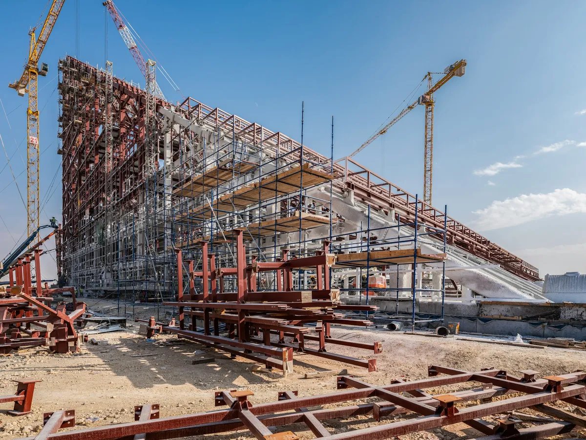 A large building rising inside steel scaffolding with two tower cranes on a clear day.