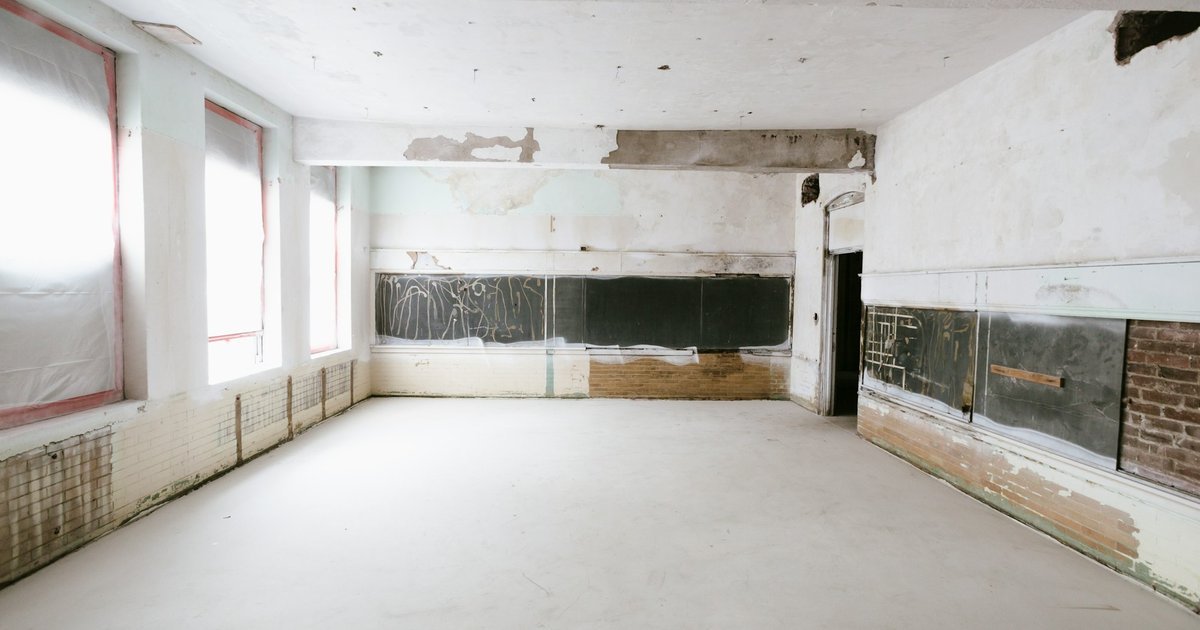 An empty room under renovation with bare plaster walls, tall windows covered in plastic on the left and a doorway at the back.