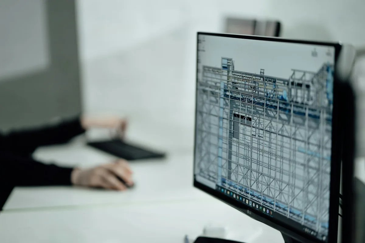 A hand on a mouse next to a monitor showing a 3D steel-frame model.